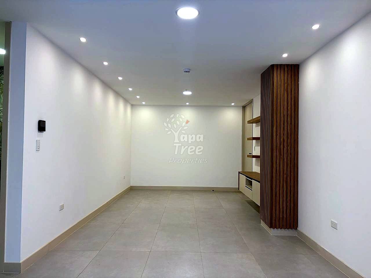 Empty living room with recessed ceiling lights, beige tile floor, white walls, and wood-slat accent wall with built-in shelving and microwave.