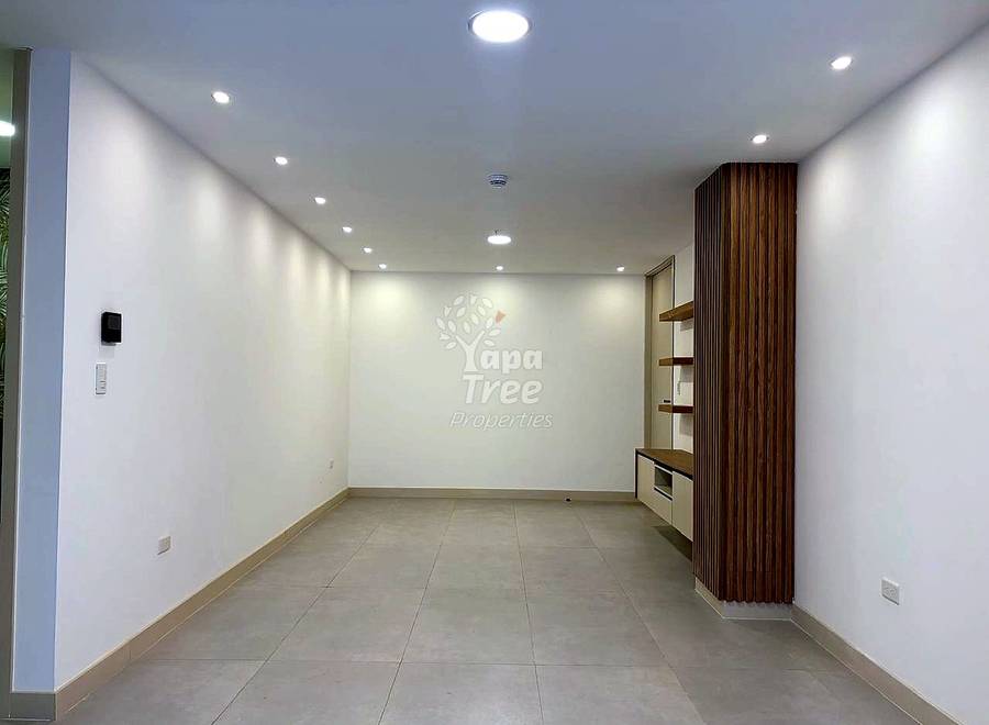 Empty living room with recessed ceiling lights, beige tile floor, white walls, and wood-slat accent wall with built-in shelving and microwave.