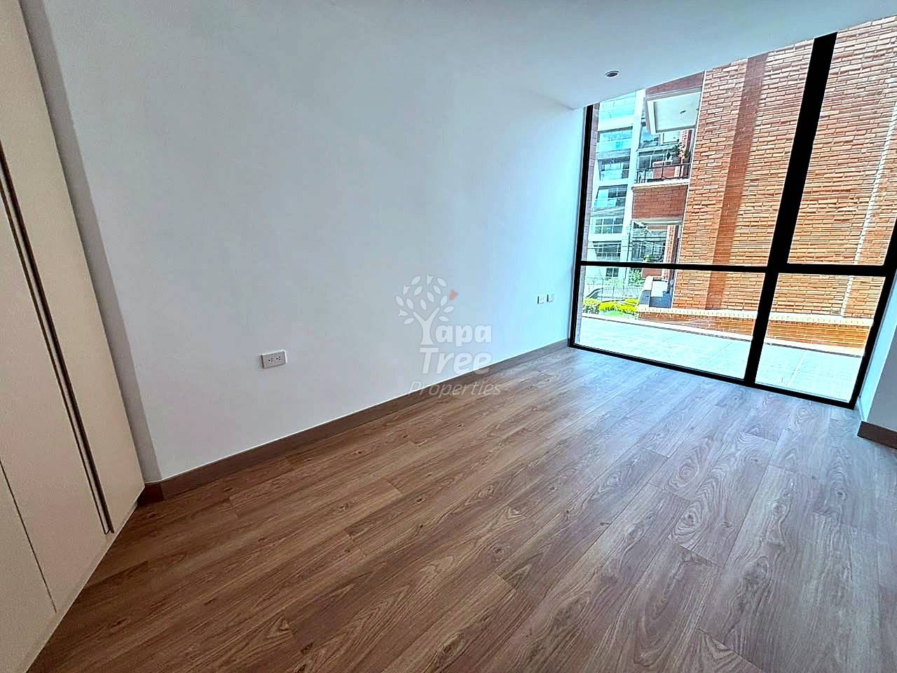 Empty room with laminate wood flooring, white walls, large window with black frame overlooking brick building, single outlet visible.