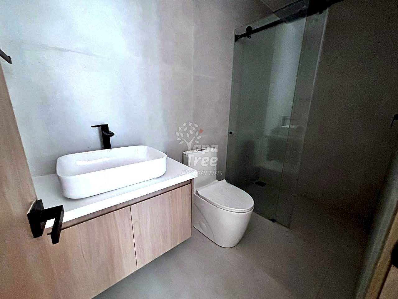 Modern bathroom with white oval vessel sink on light wood vanity, black faucet, toilet, and glass shower enclosure with black frame.