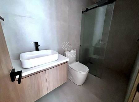 Baño moderno con lavamanos blanco ovalado sobre vanitory en madera clara, grifo negro, inodoro, y ducha con mamparas de vidrio marco negro.