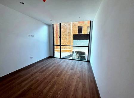 Habitación vacía con paredes blancas, piso de laminado de madera, y ventana grande con vista al patio de edificio de ladrillo con luz natural.