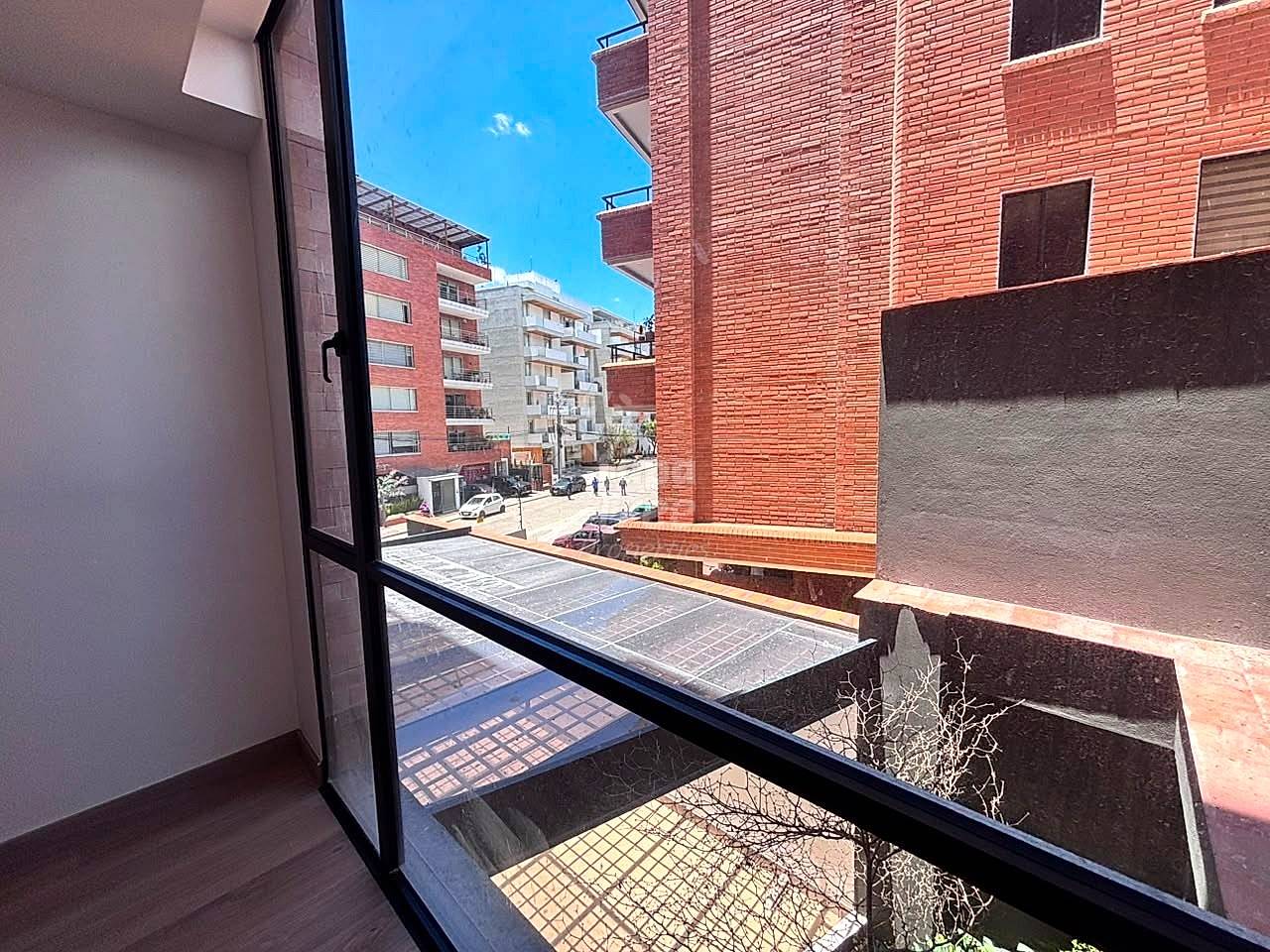 Interior view through open glass door to balcony with brick railing overlooking urban street and multi-story brick apartment buildings under clear sky