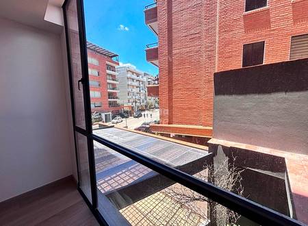 Vista interior a través de puerta de cristal abierta hacia balcón con baranda de ladrillo con vista a calle urbana y edificios de departamentos de varios pisos