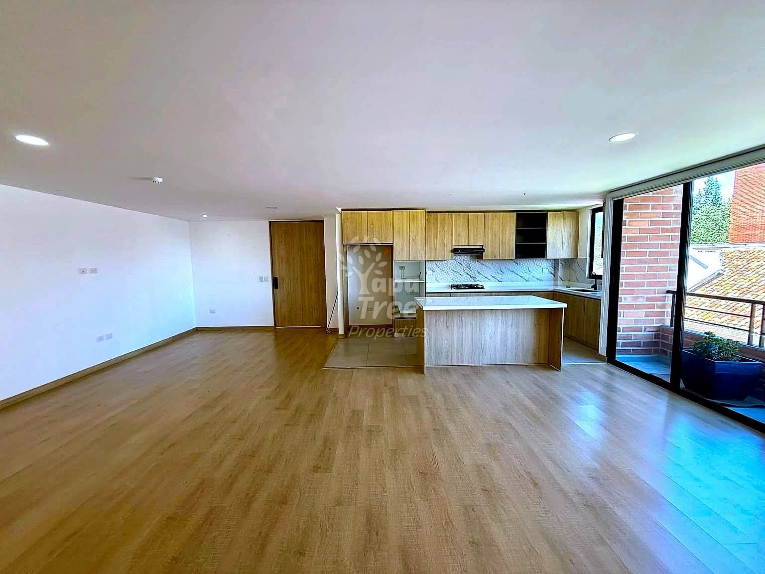 Open-plan living area with wood-tone laminate flooring, integrated kitchen with wooden cabinetry and island counter, recessed ceiling lights, and glass door access to brick terrace.