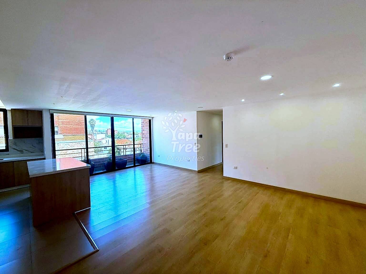 Open-plan departamento with wood flooring, brown kitchen island, recessed ceiling lights, and large glass doors opening to a balcony with city views.