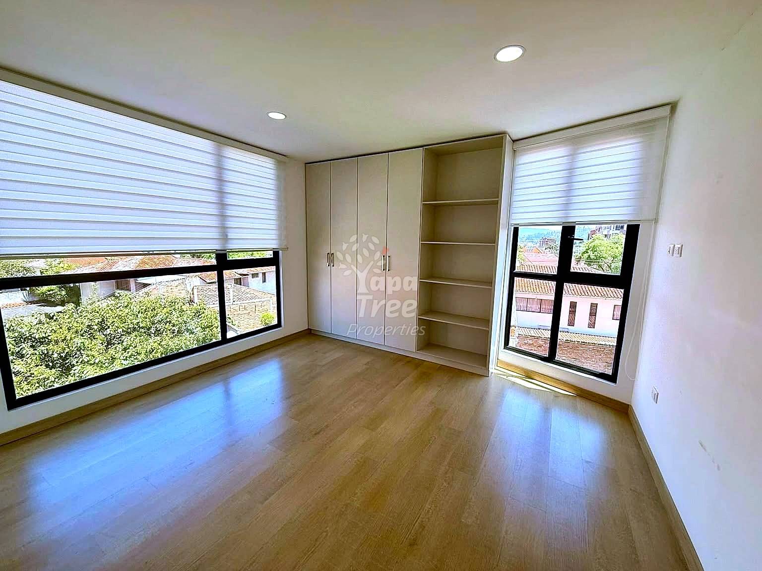 Bedroom with wooden floor, built-in wardrobe and shelving unit, two windows with roller blinds showing city views, recessed ceiling lights.
