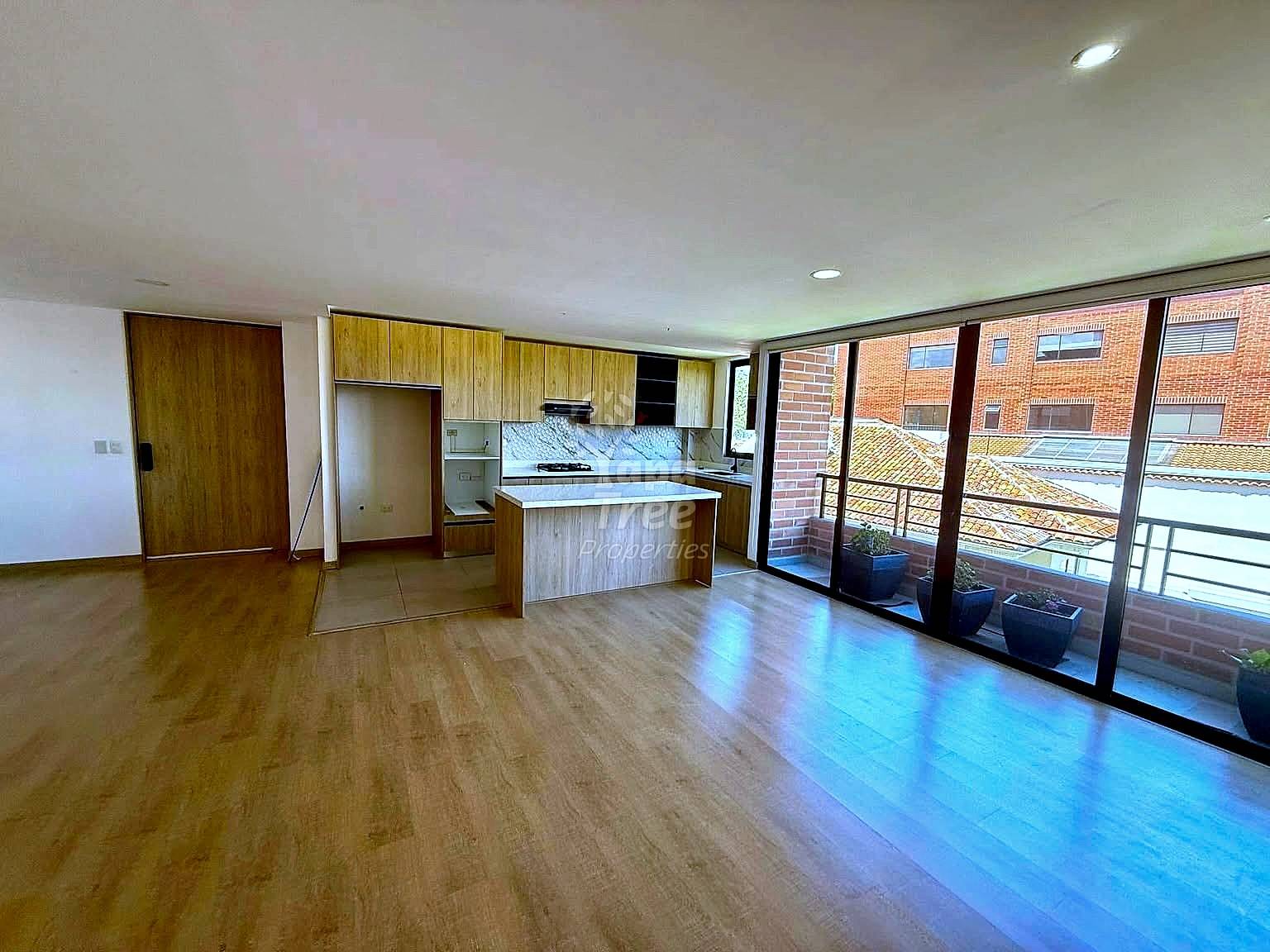 Open-plan living area with wood laminate flooring, integrated cocina with wooden cabinets and island counter, recessed ceiling lights, and glass sliding doors opening to a balcony overlooking brick buildings.