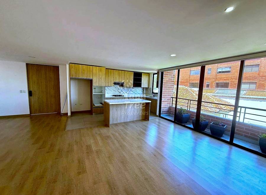 Open-plan living area with wood laminate flooring, integrated cocina with wooden cabinets and island counter, recessed ceiling lights, and glass sliding doors opening to a balcony overlooking brick buildings.