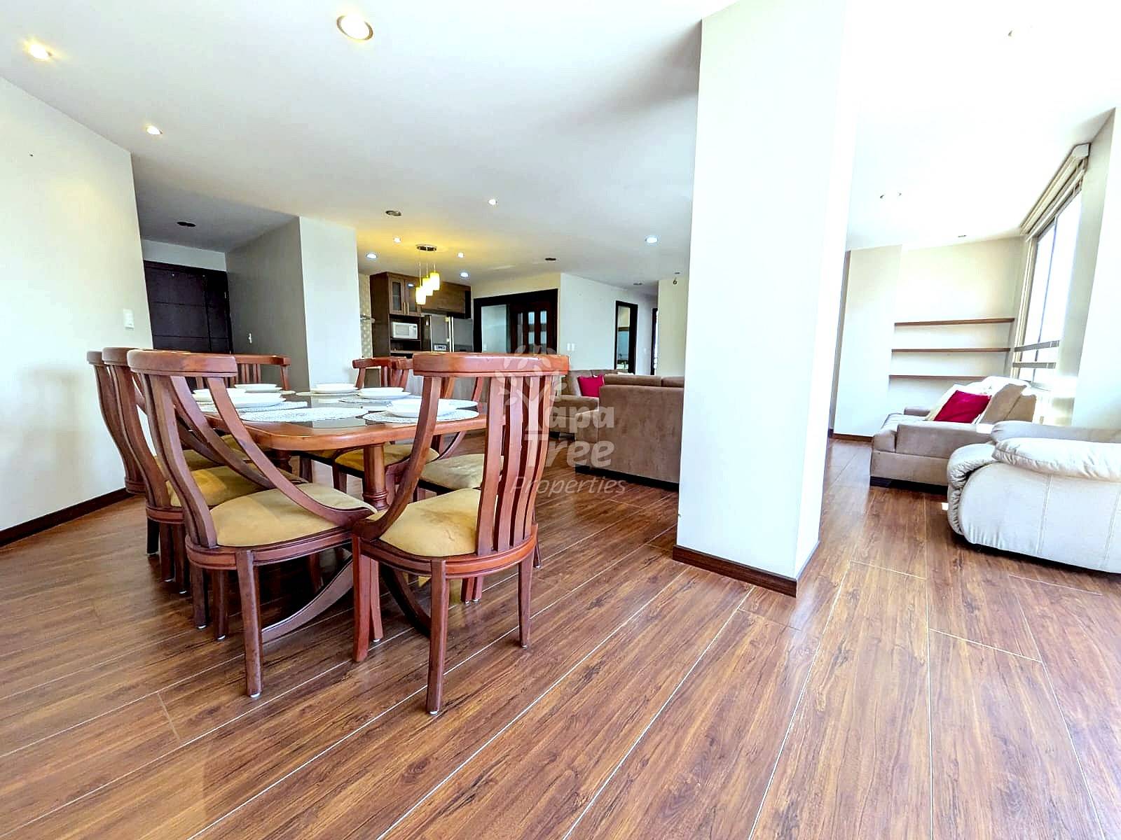 Open-concept dining and living room with wooden table, red chairs, wood flooring, and kitchen visible in background with recessed lighting.