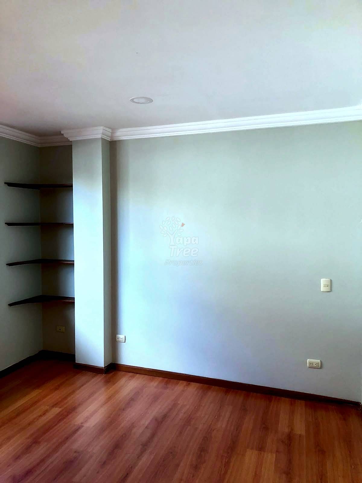 Empty room with wooden laminate flooring, cream walls, white crown molding, built-in dark wood shelves, and recessed ceiling light.