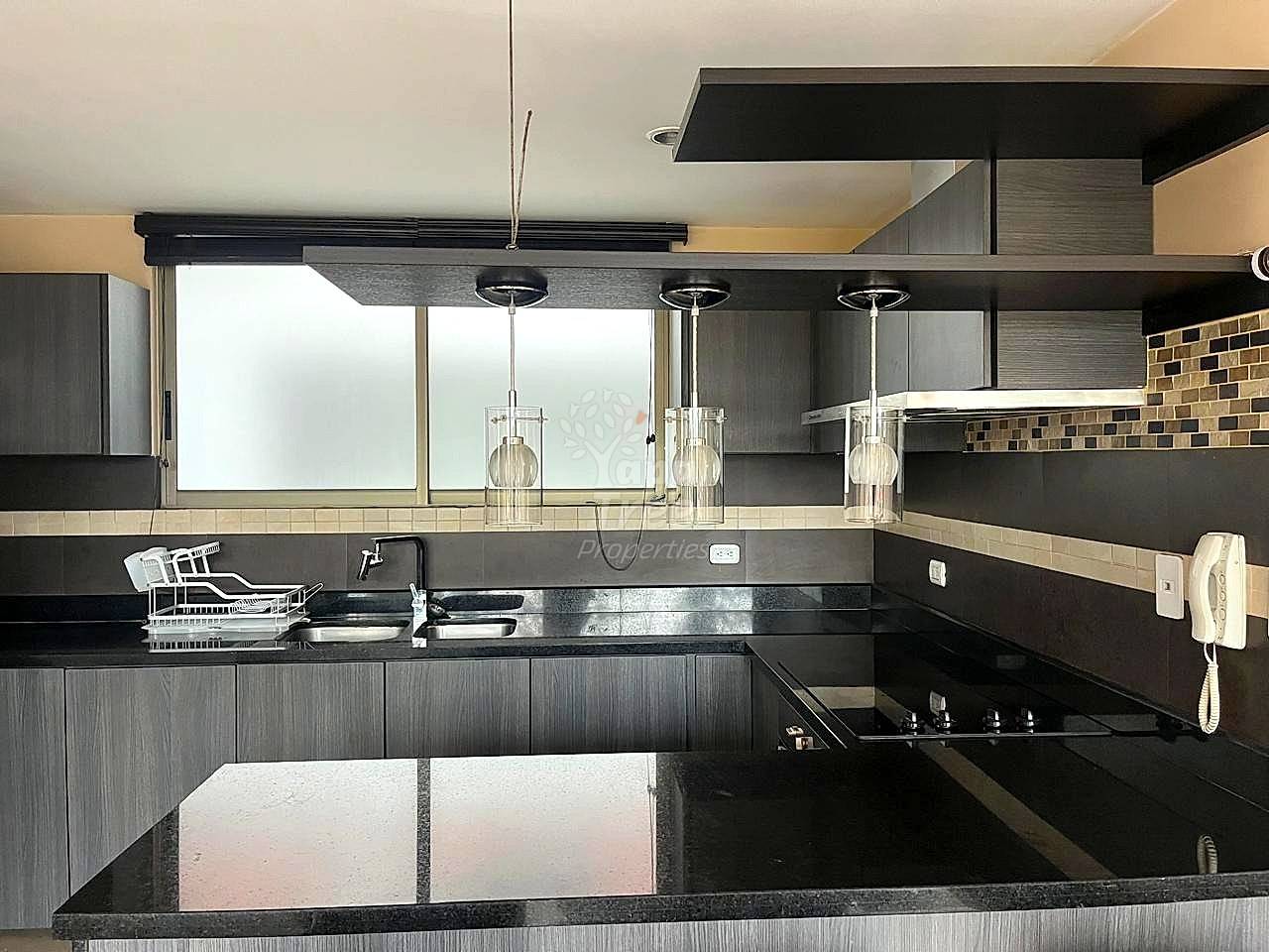 Modern cocina with dark gray cabinetry, black granite countertop, stainless steel sink, and three pendant lights over counter. Tile backsplash visible.