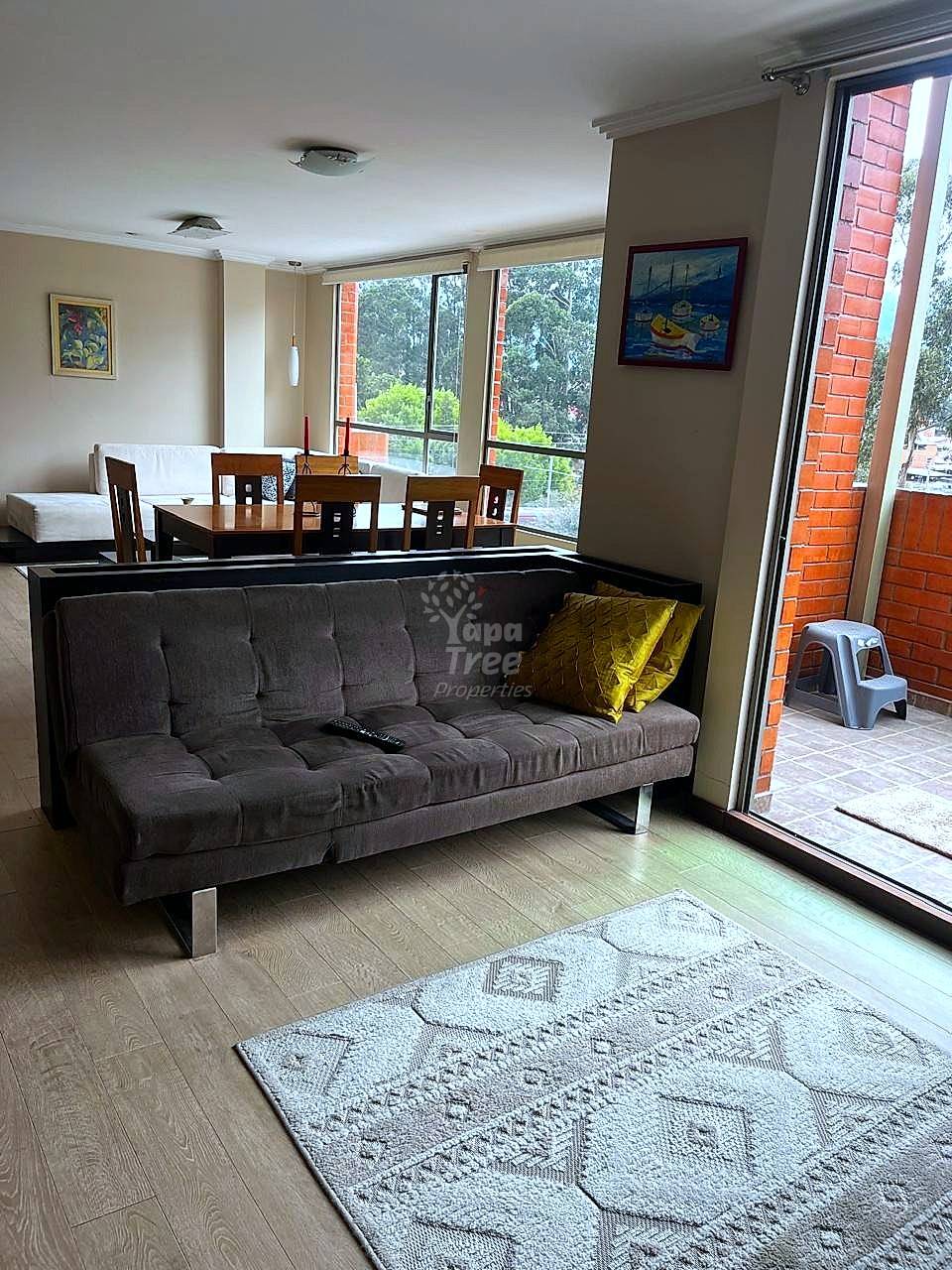 Open living room with gray upholstered sofa, wooden dining table with chairs, large windows overlooking trees, wood flooring, and brick exterior wall visible through glass door.