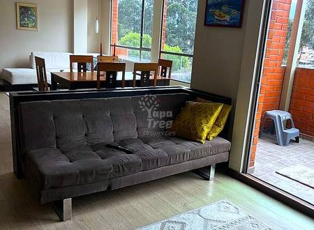 Sala abierta con sofá tapizado gris, mesa de comedor de madera con sillas, grandes ventanas con vista a árboles, piso de madera, y pared de ladrillo visible a través de puerta de vidrio.