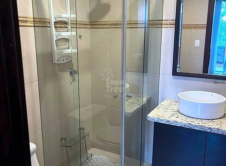 Baño moderno con ducha de cristal, regadera de lluvia, lavamanos blanco sobre vanidad oscura con mesada de granito, e inodoro blanco.