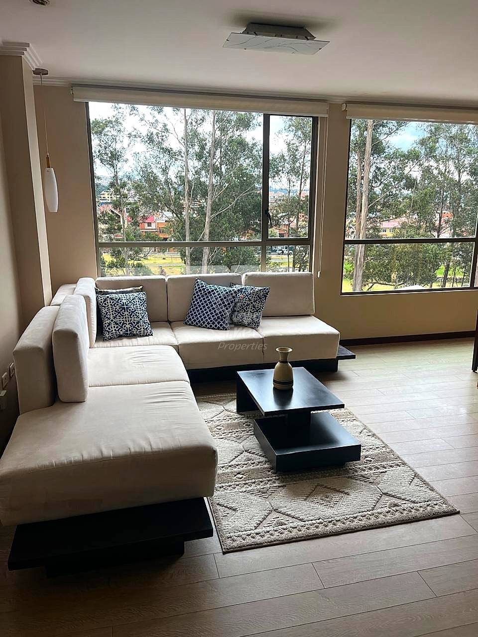 Living room with beige L-shaped sectional sofa, black coffee table, patterned pillows, and large windows overlooking green landscape and Cuenca cityscape.