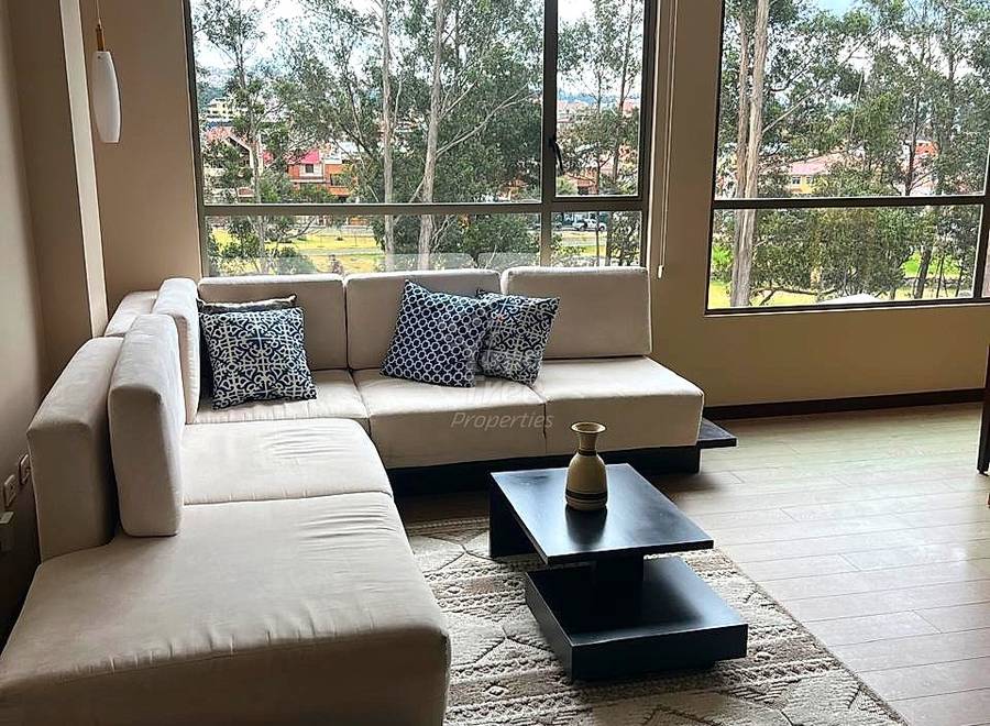 Sala con sofá seccional beige, mesa de centro negra, cojines estampados y ventanales amplios con vista al paisaje verde y ciudad de Cuenca.