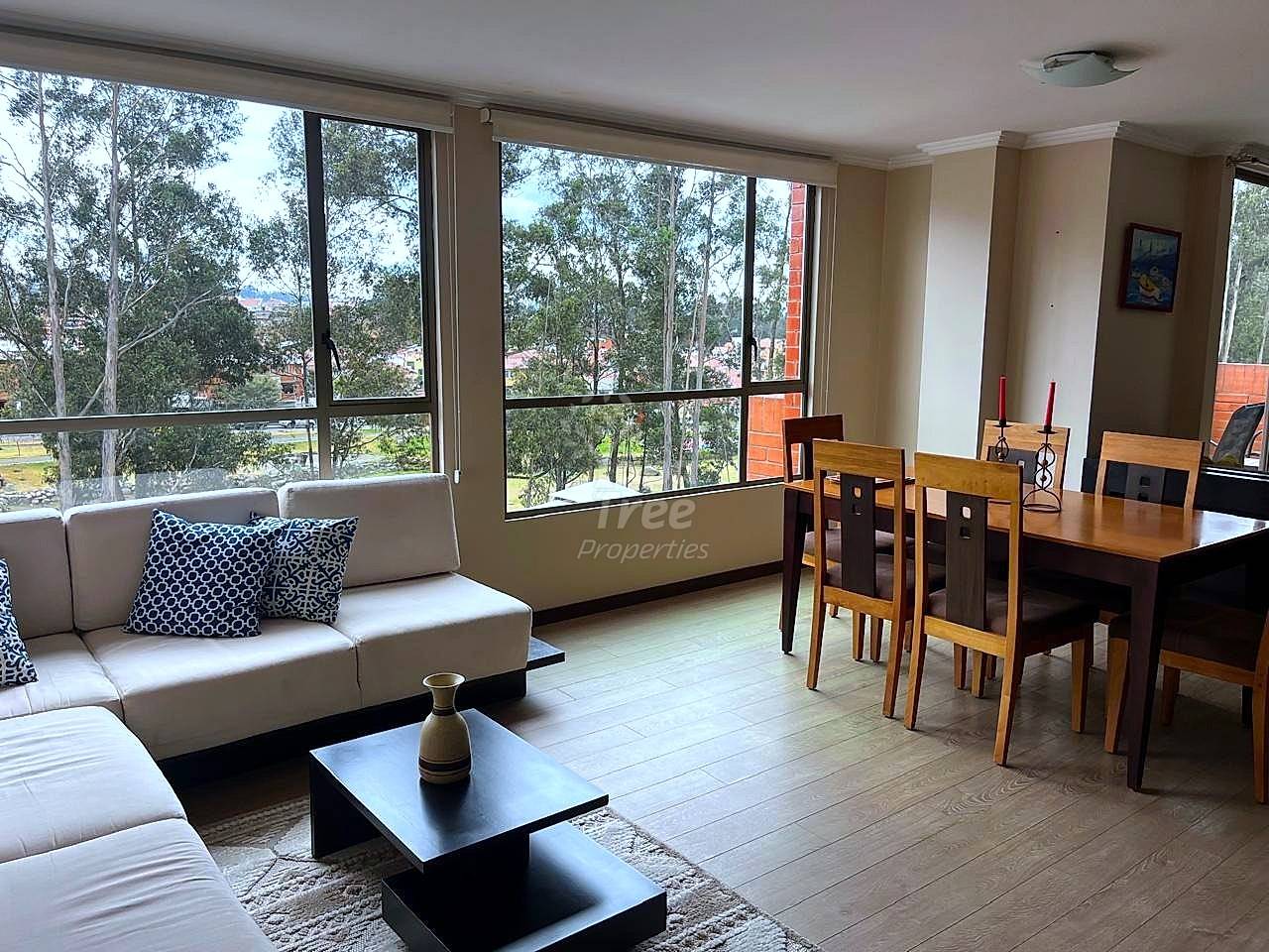 Sala y comedor abiertos con sofá beige, mesa de centro negra, mesa de comedor de madera con sillas, ventanas grandes con vista a árboles y paisaje, piso de madera clara.