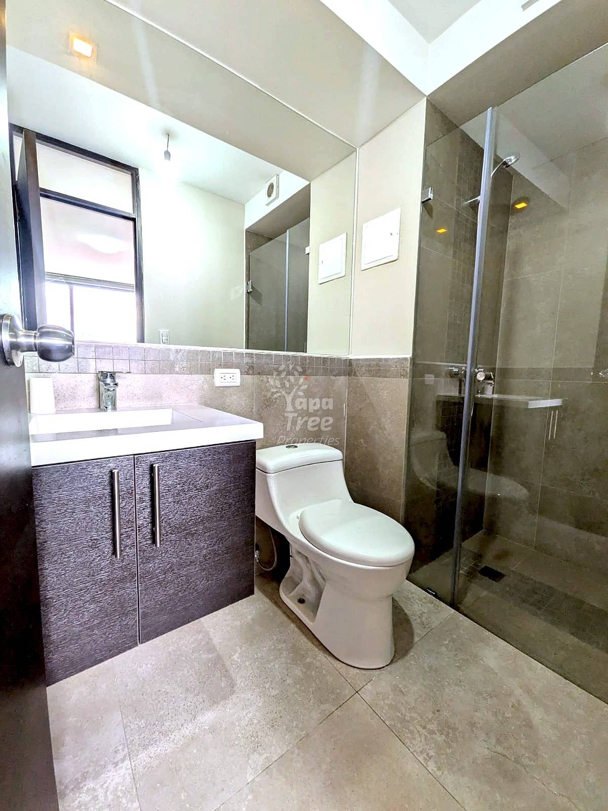 Modern bathroom with tile flooring, dark vanity cabinet, white sink, toilet, glass shower enclosure, and natural light from window.