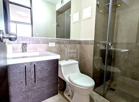 Modern bathroom with tile flooring, dark vanity cabinet, white sink, toilet, glass shower enclosure, and natural light from window.