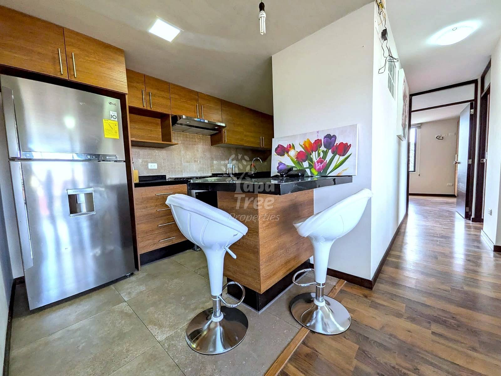 A modern kitchen with stainless steel refrigerator, wooden cabinets, and a breakfast bar with two white stools.