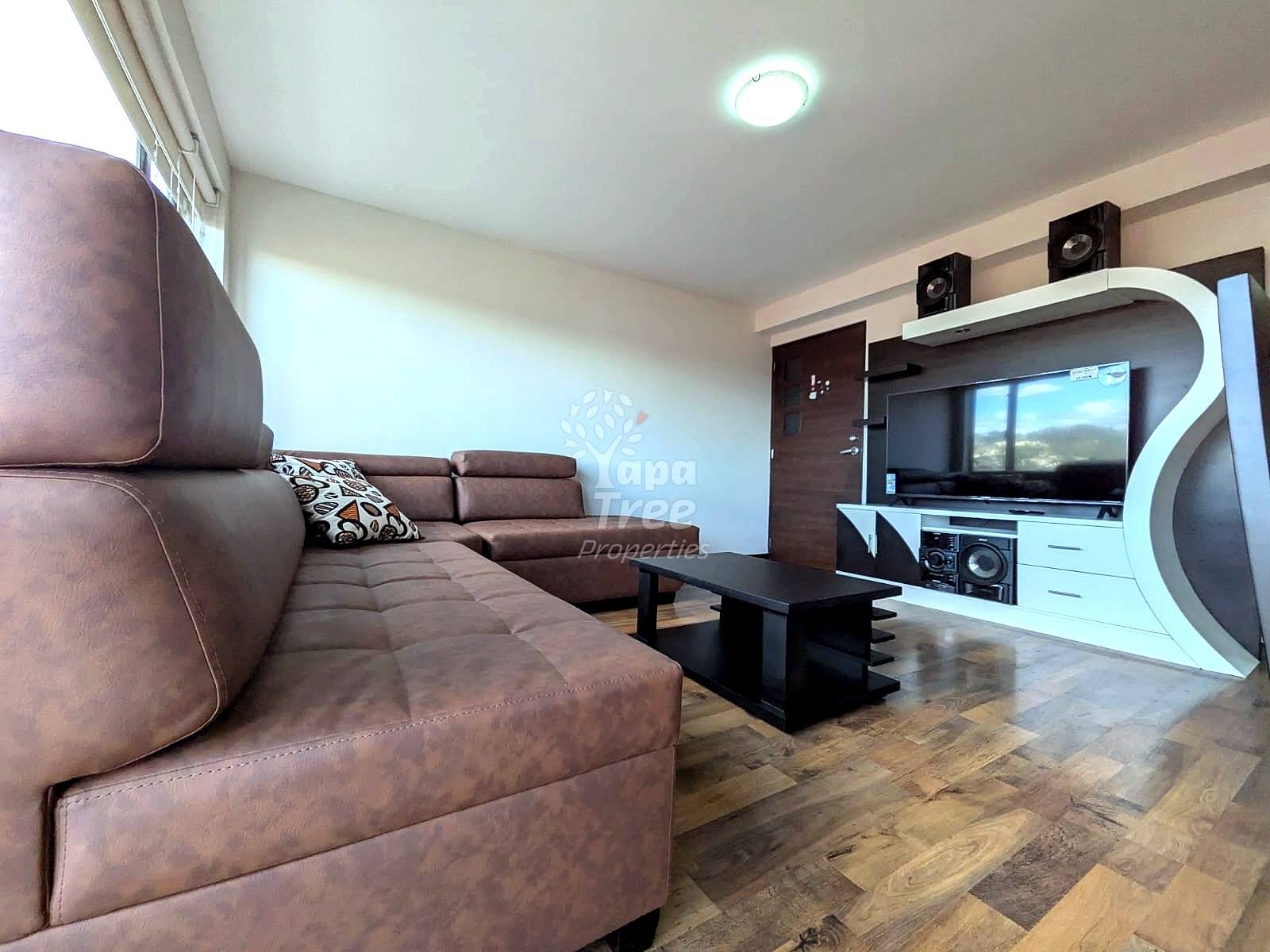 Living room with brown leather sectional sofa, black coffee table, wall-mounted TV with white entertainment unit, recessed ceiling light, and wood laminate flooring.