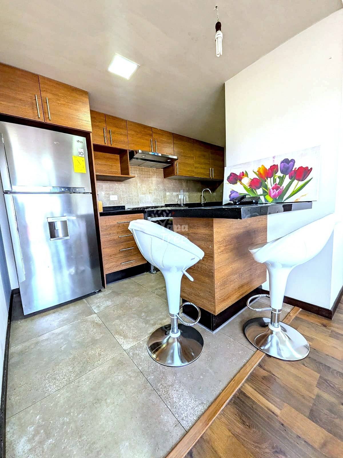 Modern kitchen with wood cabinetry, stainless steel refrigerator, black countertop, two white bar stools, tile and wood flooring, and recessed lighting.