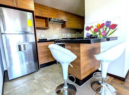 Modern kitchen with wood cabinetry, stainless steel refrigerator, black countertop, two white bar stools, tile and wood flooring, and recessed lighting.