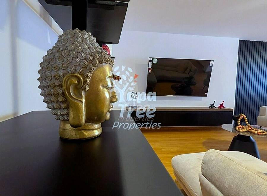 Living room with wooden floor, black dining table, beige sofa, wall-mounted TV, and gold Buddha sculpture decoration.