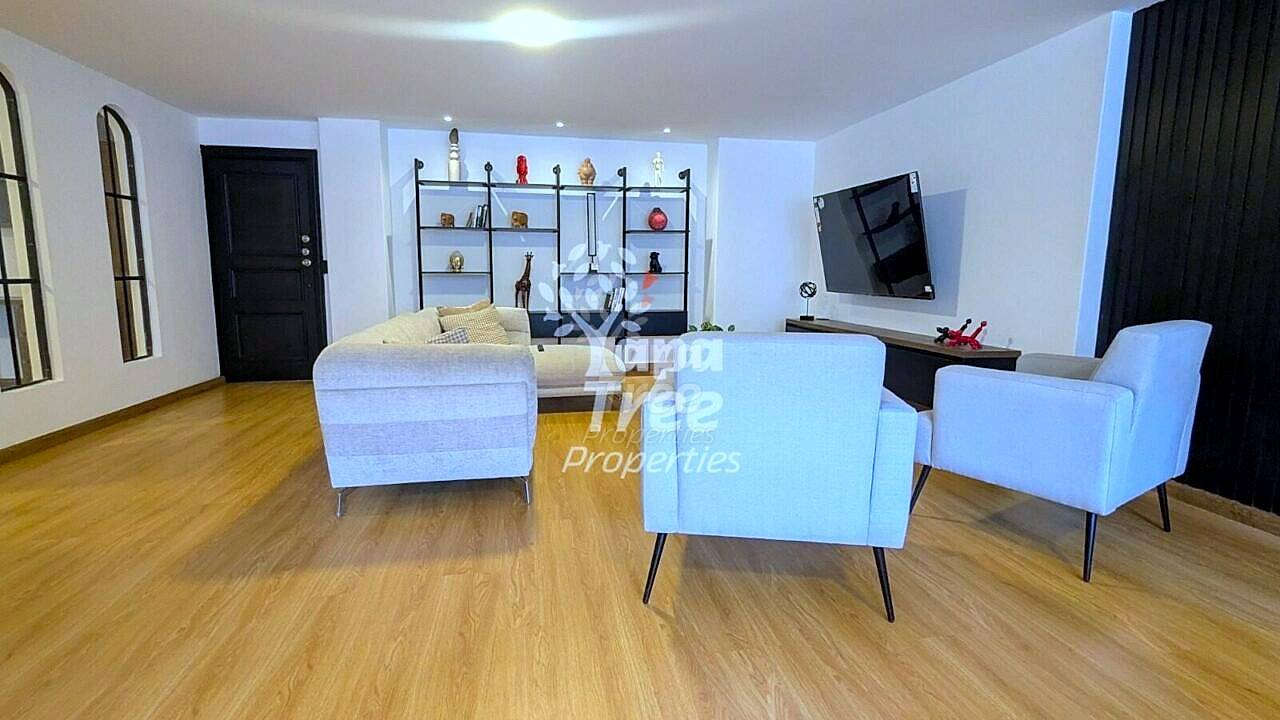 Open-plan living room with wooden flooring, white upholstered armchairs, beige sofa, wall-mounted TV, black shelving unit with decorative items, and recessed ceiling lights.