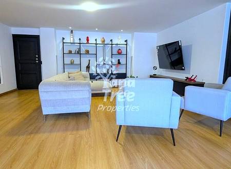 Open-plan living room with wooden flooring, white upholstered armchairs, beige sofa, wall-mounted TV, black shelving unit with decorative items, and recessed ceiling lights.