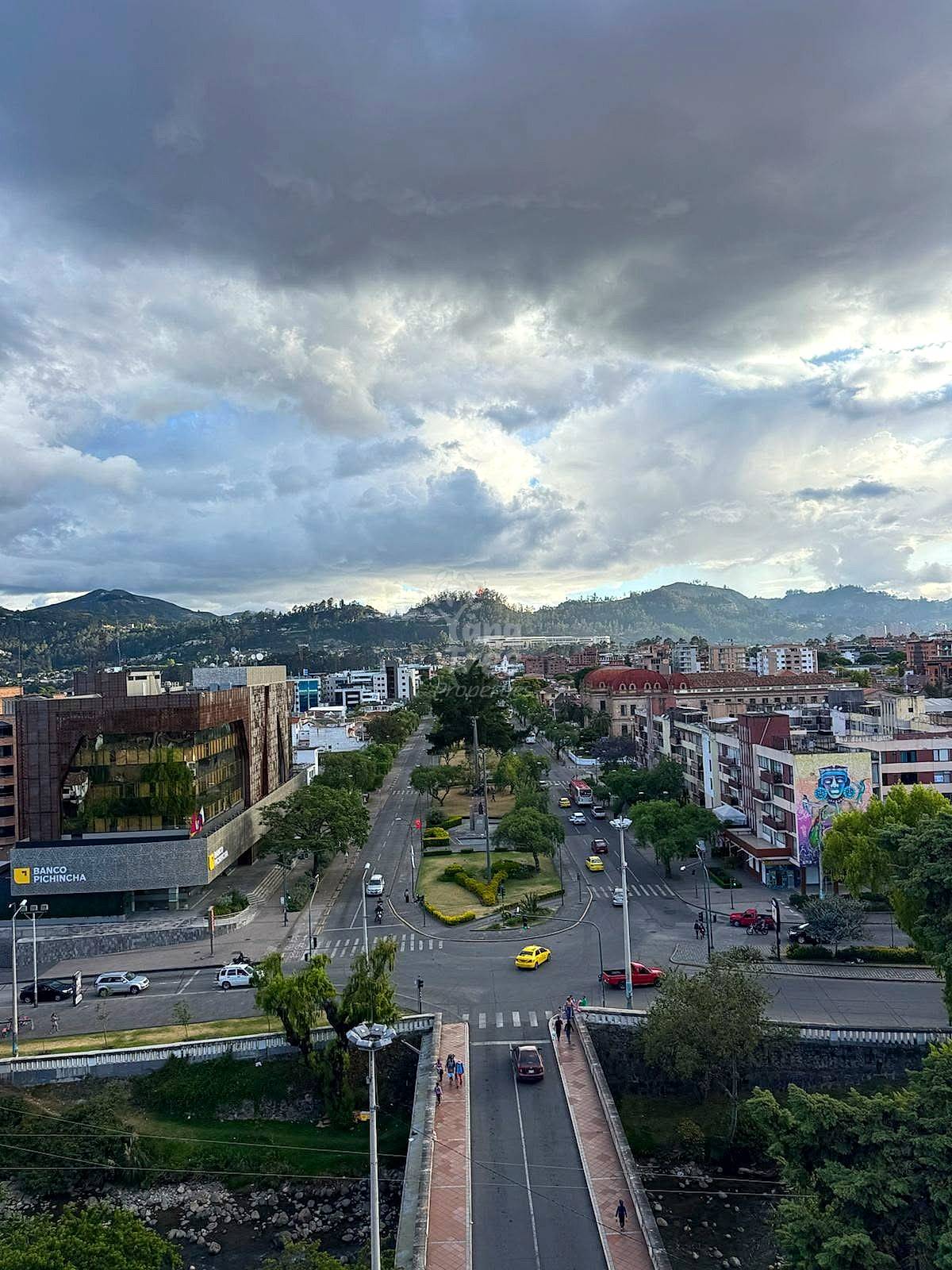 Elevated view of Cuenca city center with commercial buildings, streets, green plaza, mountains, and overcast sky.