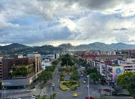 Elevated view of Cuenca city center with commercial buildings, streets, green plaza, mountains, and overcast sky.