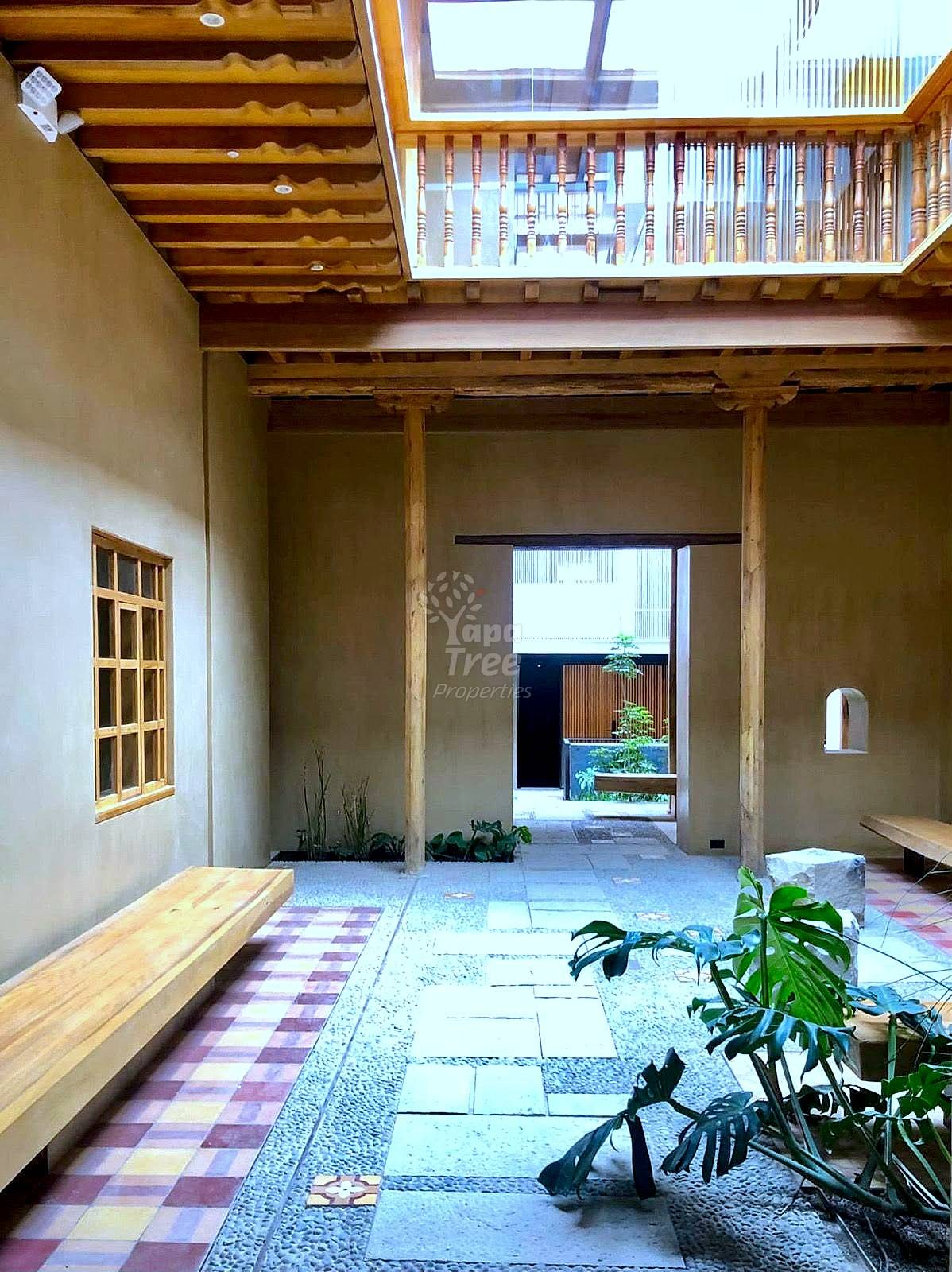 Interior courtyard with exposed wooden beams, tile flooring, wooden bench, potted plants, and doorway opening to garden area.