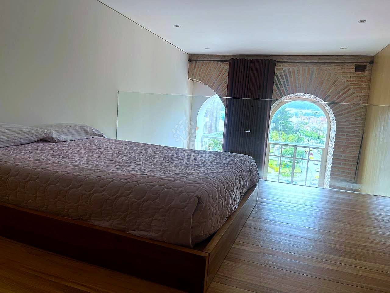 Bedroom with brown platform bed, wooden flooring, arched window with city view, brick wall, and dark curtains.