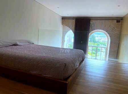 Bedroom with brown platform bed, wooden flooring, arched window with city view, brick wall, and dark curtains.