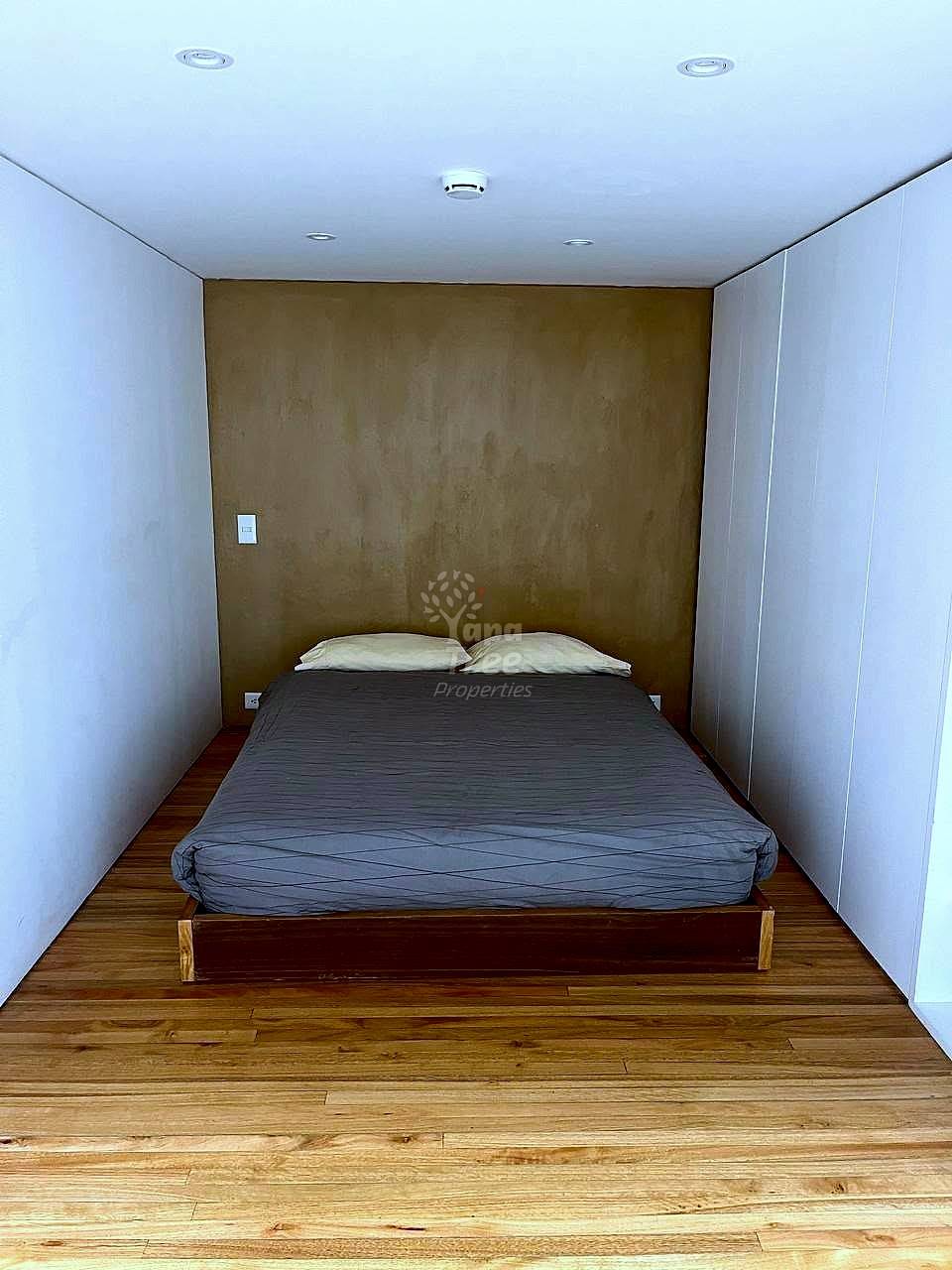 Bedroom with wooden bed, gray bedding, cream pillows, brown accent wall, white side walls, wooden floor, and recessed ceiling lights.