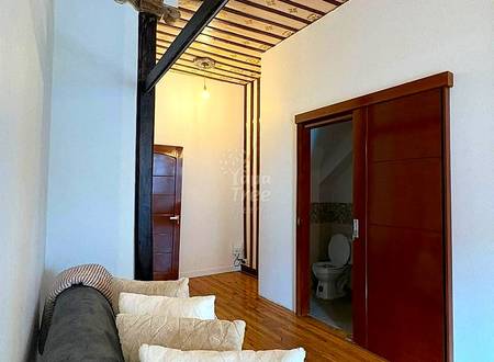 Studio apartment with wooden beam ceiling, gray sofa with white pillows, wood flooring, brown wood doors, and bathroom visible on right.