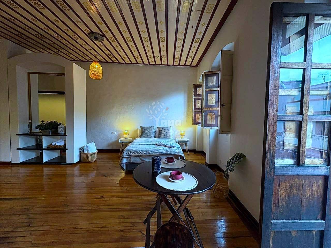 Studio suite with wooden slatted ceiling, bed with side tables, round black dining table, wooden floors, and large gridded window with city views.