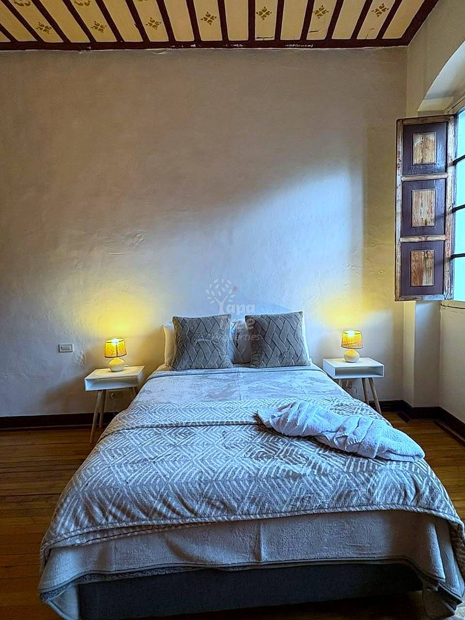 Bedroom with queen bed, patterned bedding, two wooden nightstands with table lamps, decorative wooden ceiling, and window with wooden shutters.