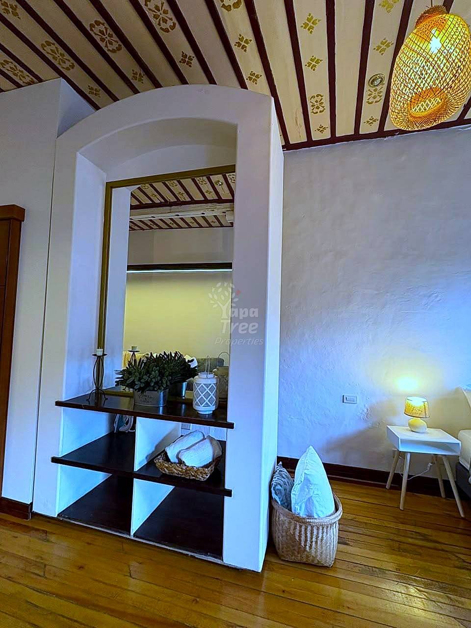 Bedroom with arched alcove shelving unit, wooden nightstand with lamp, woven basket, and decorative ceiling with patterned fabric and woven pendant light.