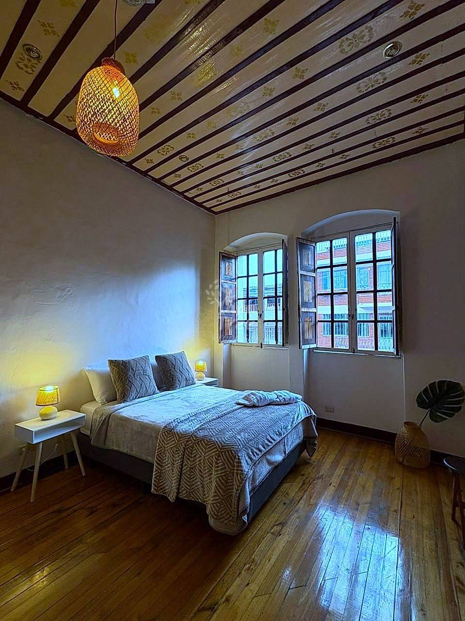 Bedroom with wooden beam ceiling, woven pendant light, bed with patterned bedding, two wooden-framed windows with city views, and wood flooring.