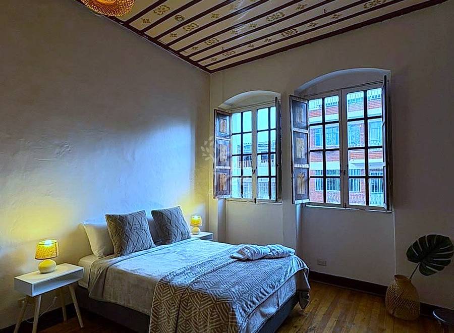 Bedroom with wooden beam ceiling, woven pendant light, bed with patterned bedding, two wooden-framed windows with city views, and wood flooring.