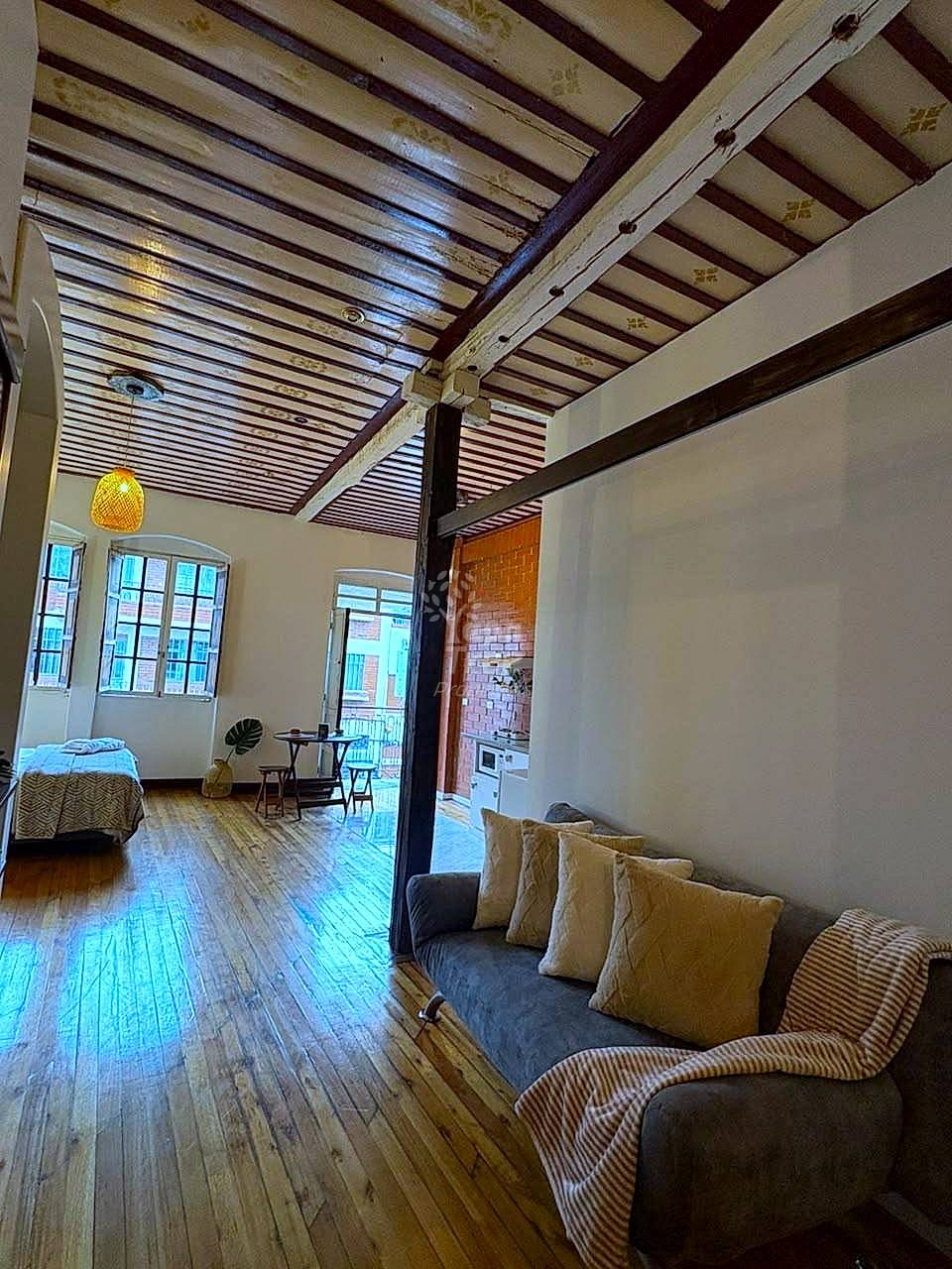 Open-plan suit with wooden ceiling beams, armchair with pillows, bed, wood flooring, windows, and brick cocina visible in background.
