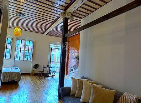 Open-plan suit with wooden ceiling beams, armchair with pillows, bed, wood flooring, windows, and brick cocina visible in background.