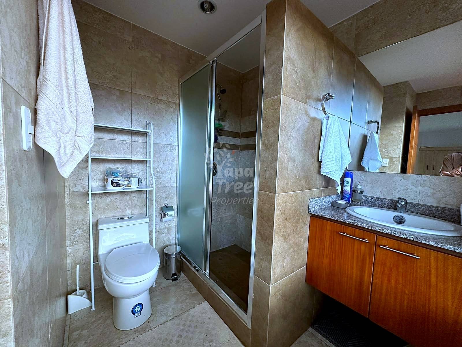 Bathroom with beige tile walls, glass-enclosed shower stall, white toilet with metal storage rack, and wooden vanity with granite countertop and mirror.