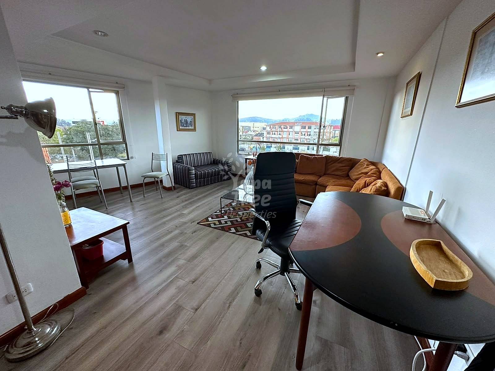 Open living area with gray laminate flooring, brown sofa, black office chair, curved desk, and large windows overlooking Cuenca city and mountains.