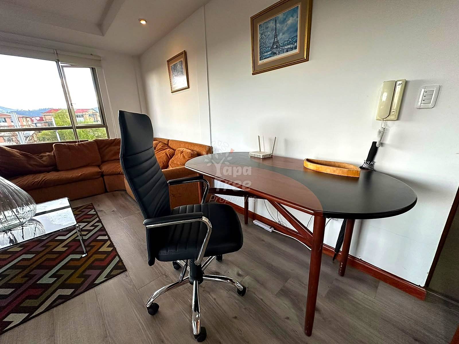 Office area with oval desk, black executive chair, brown leather sofa, framed artwork, window with city views, and wood flooring.