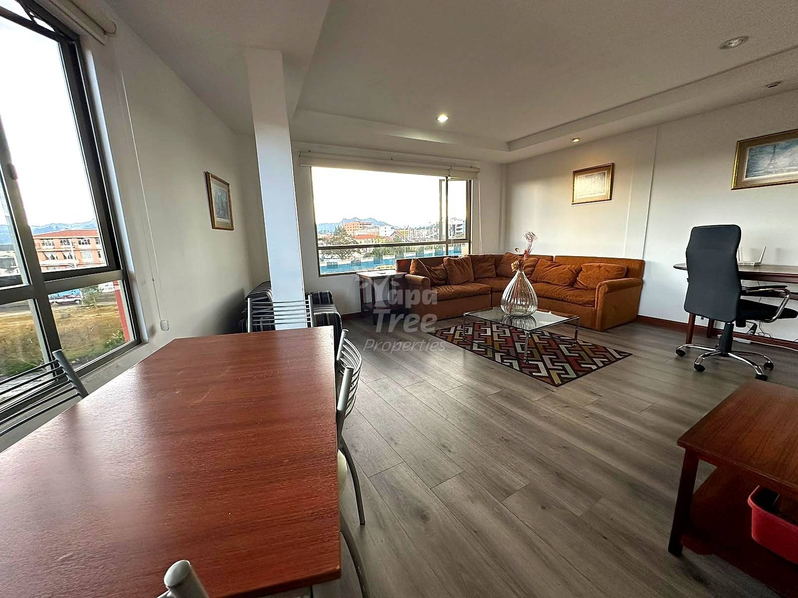 Open-plan living and dining area with brown leather sofa, wooden dining table, gray wood flooring, large windows with city views, and recessed ceiling lights.