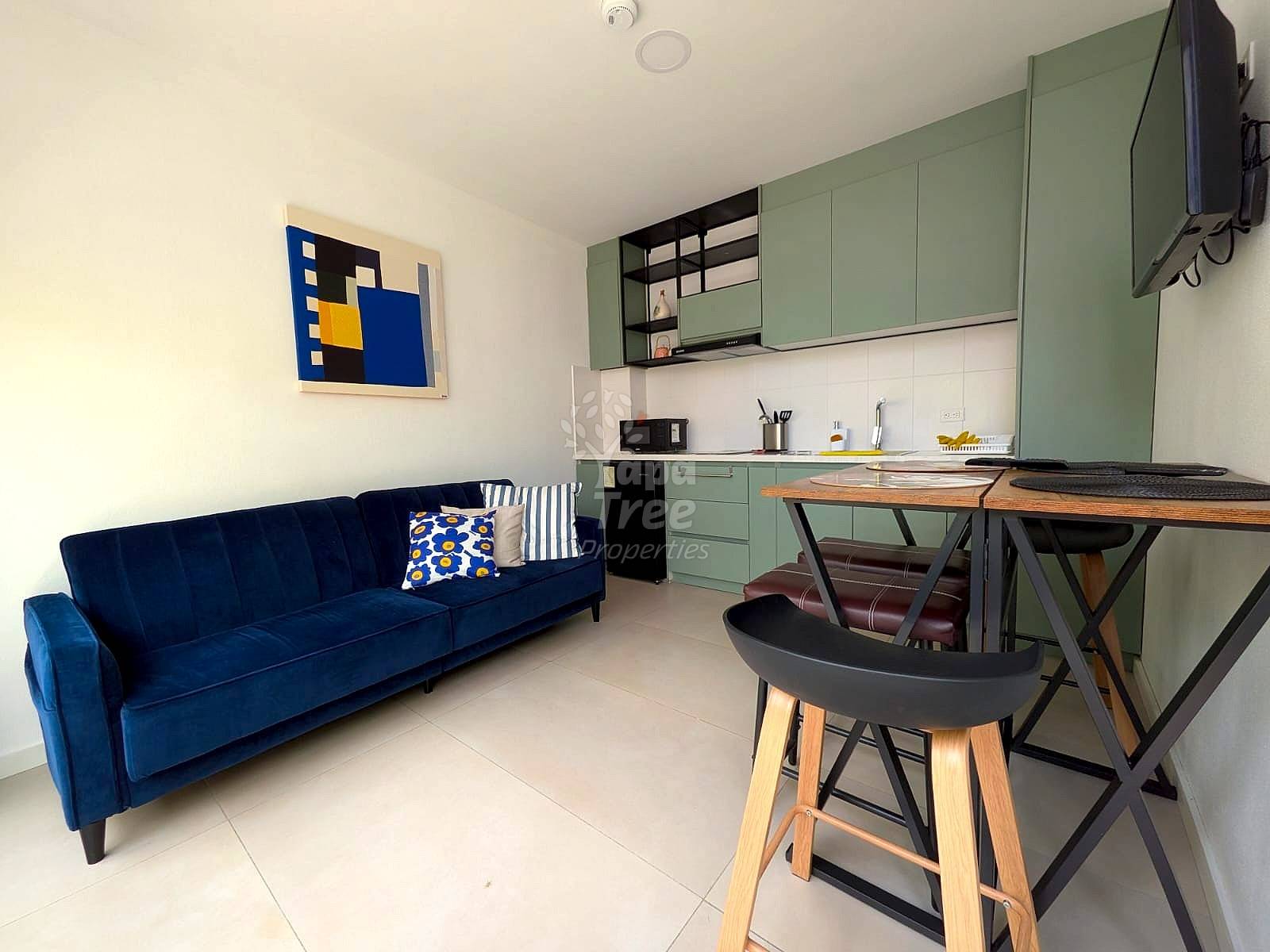 Open-concept departamento with blue sofa, dining table with black stools, green cabinetry cocina, white walls, and tile flooring.